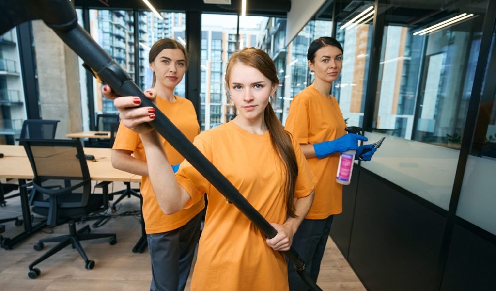 Three cleaning company workers in uniform cleaning and disinfecting office space Three cleaning company workers in uniform and protective gloves cleaning and disinfecting office space with vacuum cleaner, rag and spray
